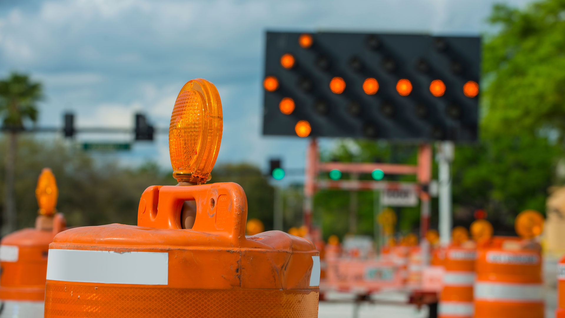 Orange traffic barrels a road under construction with an orange-lighted directional arrow in the background pointing left.